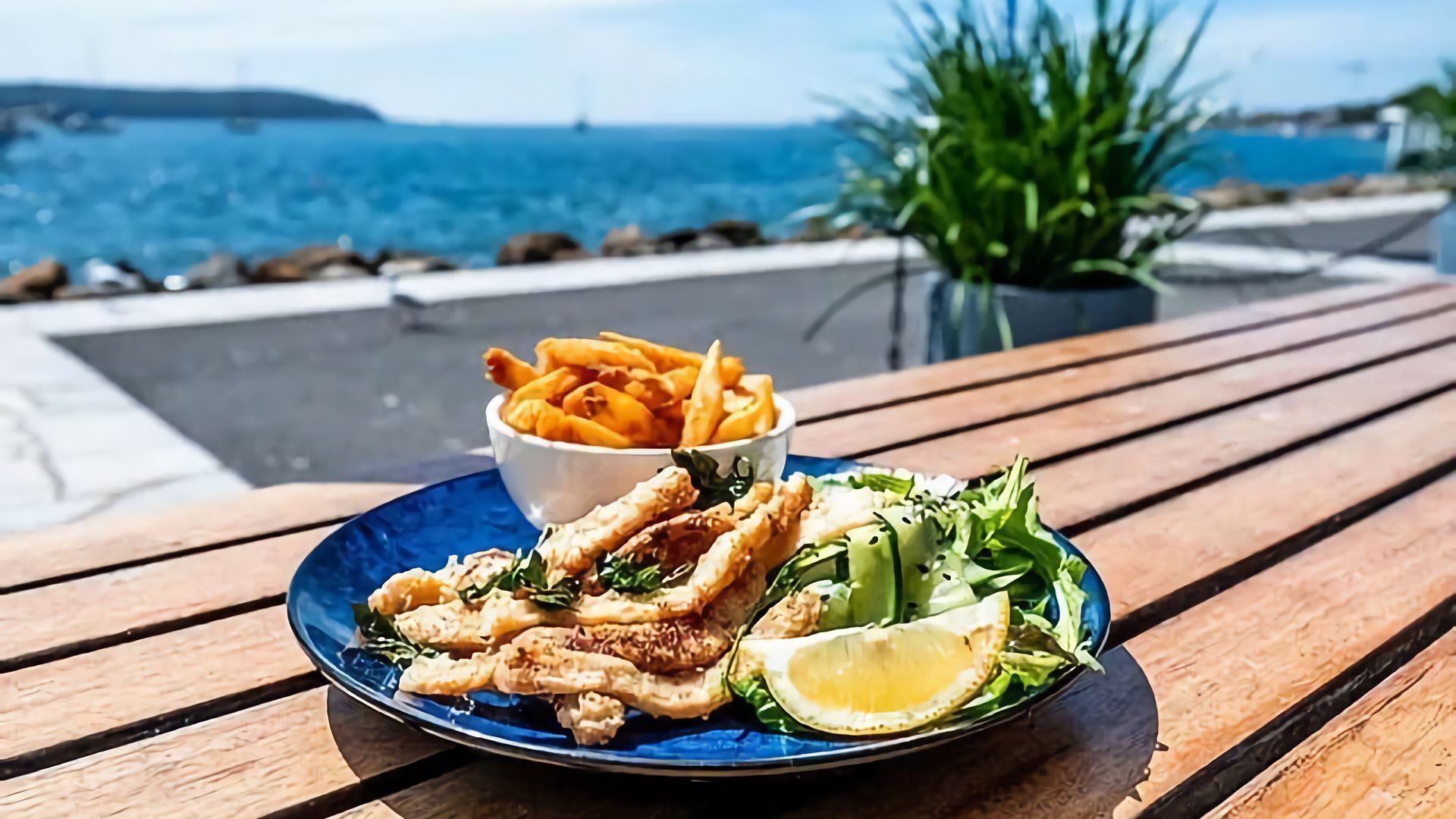 A meal of fish, fries, and salad on a wooden table with a view of the sea.