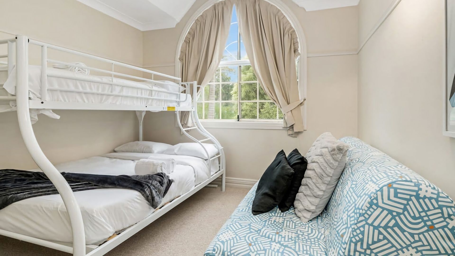Een stapelbed met een wit frame, een enkel bed met een beddengoed met een patroon, en een raam met gordijnen.