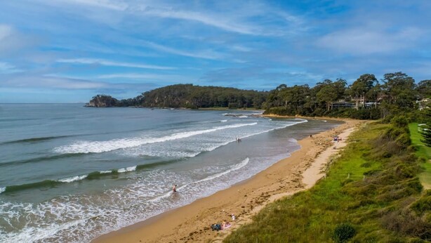 A coastal beach with waves, a sandy shore, and a forested hill in the background.