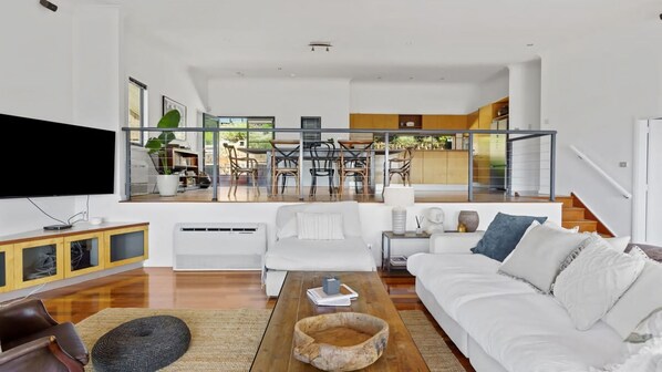 A modern living room with a TV, white sofas, a wooden coffee table, and a view of a dining area through large windows. - Hilltop Hideaway Merewether Family Escape (Merewether)
