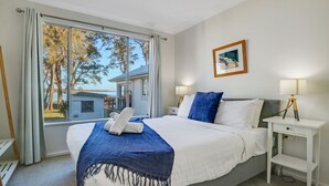 A bedroom with a large window, a bed with white and blue bedding, two bedside tables with lamps, and a framed picture on the wall. - Lake Front Home Relax Unwind (Budgewoi)