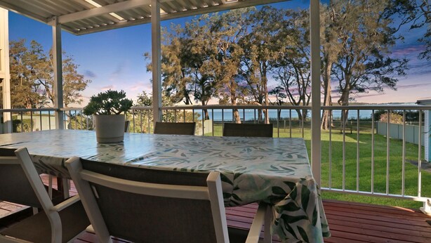 An outdoor patio with a table and chairs, a potted plant, and a view of a grassy area and trees. - Lake Front Home Relax Unwind (Budgewoi)