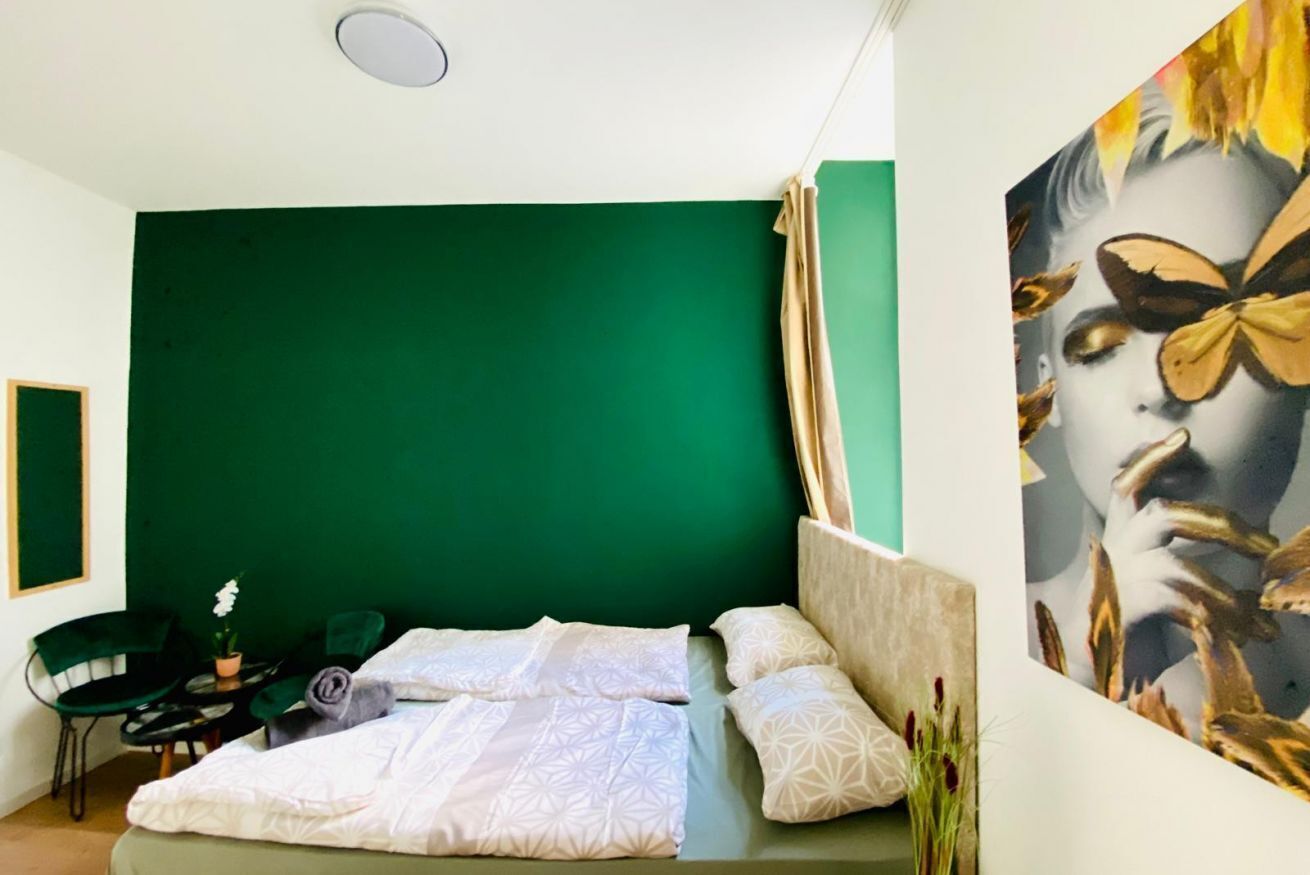 Ein Zimmer mit grüner Akzentwand, einem Bett mit gemusterten Betttextilien, einem Sessel, einem kleinen Tisch mit Pflanze und einem Gemälde an der Wand.