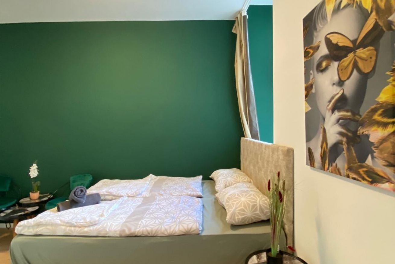 A hotel room with a green wall, a bed with patterned bedding, a small table with a plant, and a painting on the wall.