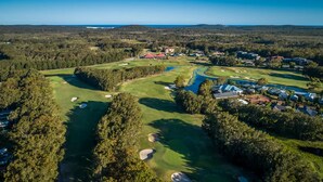An aerial view of a golf course with bunkers, a water hazard, and surrounding residential buildings. - Hitters Paradise Stay Play (Salamander Bay)