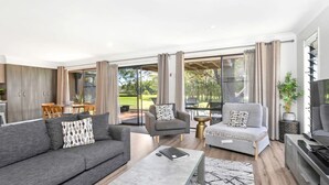 A modern living room with a grey sofa, armchair, and coffee table. There is a dining area with a table and chairs, and a TV on a stand. Large windows offer a view of the outdoors. - Hitters Paradise Stay Play (Salamander Bay)