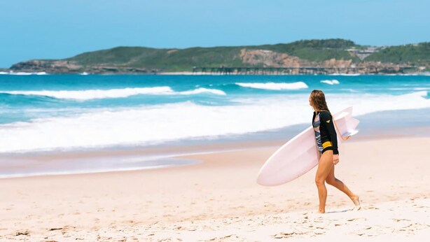 Una persona camina sobre una platja sorrenca amb una taula de surf, ones oceàniques i un turó verd a la distància.