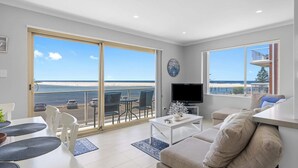 Ein großzügiger Wohnraum mit Balkonblick auf den Strand, einem Flachbildfernseher und einem Essbereich mit Tisch und Stühlen.