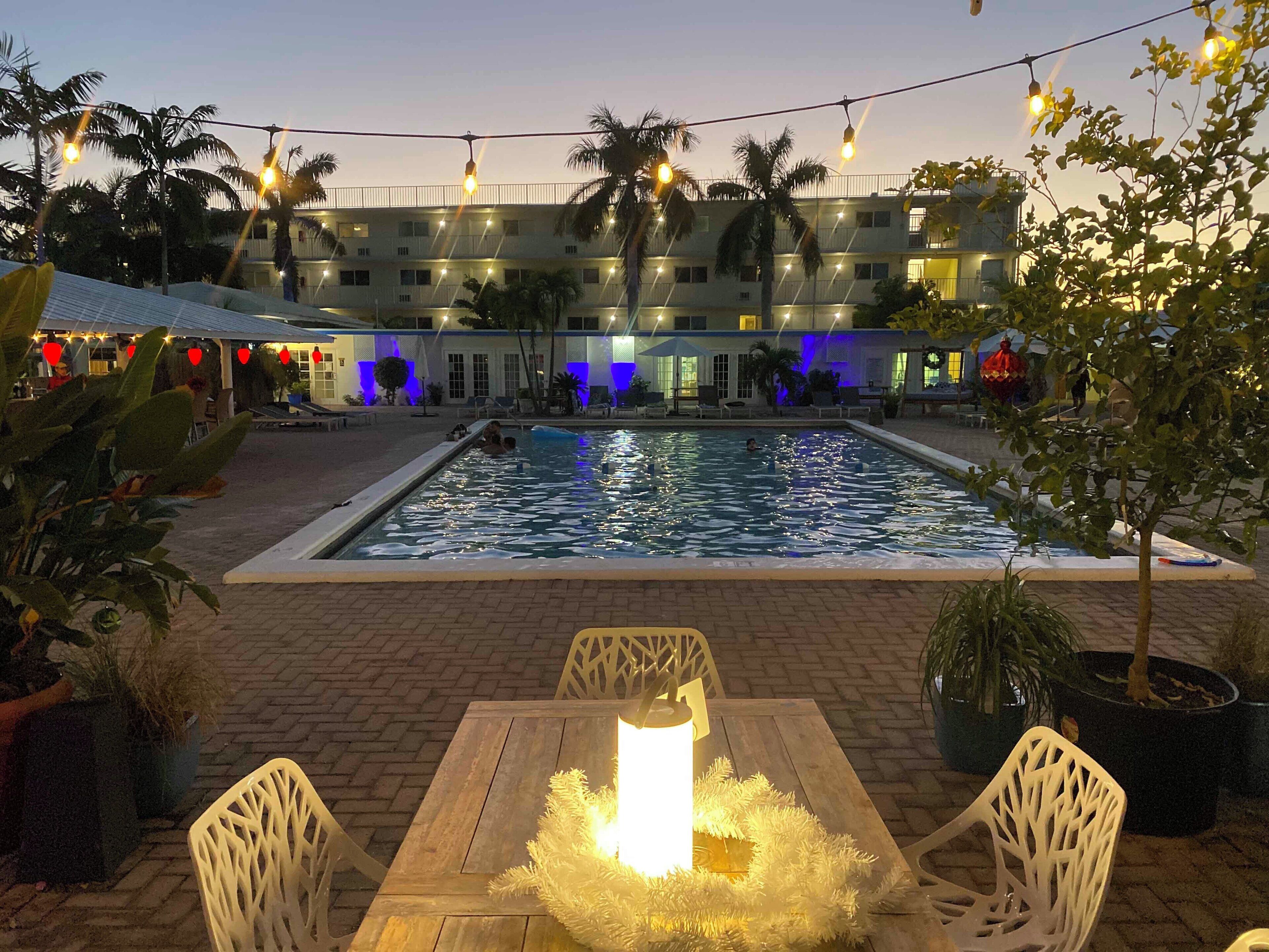 A swimming pool with a wooden table and chairs in the foreground, surrounded by potted plants.