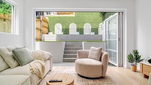 Una sala de estar moderna con un sofá, un sillón y una mesa de centro. Una escalera conduce a un balcón con un muro verde y ventanas blancas.