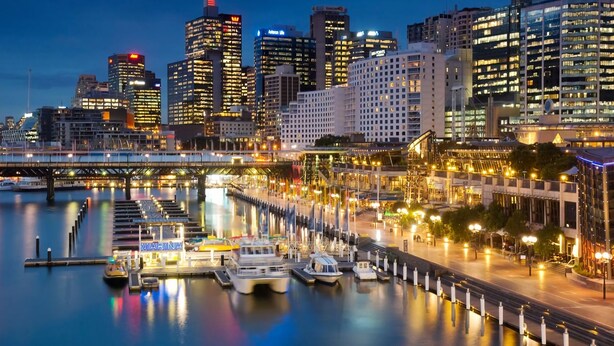 Una marina con barcos amarrados, un puente y un horizonte urbano con edificios iluminados al anochecer.