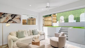 Una sala moderna con un sofá blanco, una mesa de centro redonda y un sillón beige. Un ventanal ofrece vistas a un jardín verde y una cerca de madera.
