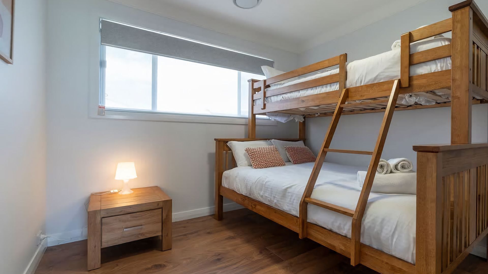 A bunk bed room with a wooden floor, a nightstand with a lamp, and a window with blinds.