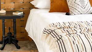 A neatly made bed with a white comforter and a striped blanket. There is a wooden headboard, a small bedside table with a book, and a single orange pillow. - Coast and Roast - Sunny Shell Cove Escape (Shell Cove)