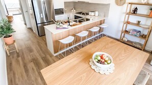 A modern kitchen with a central island, bar stools, and a dining table with a fruit bowl. - Coast and Roast - Sunny Shell Cove Escape (Shell Cove)