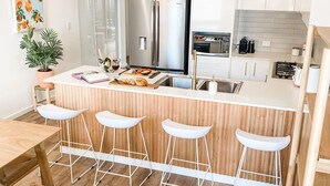 A modern kitchen with a central island, bar stools, stainless steel appliances, and a potted plant. - Coast and Roast - Sunny Shell Cove Escape (Shell Cove)
