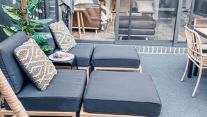 A modern outdoor seating area with a sofa and ottomans, a small table with a plate of food, and a wicker chair. - Coast and Roast - Sunny Shell Cove Escape (Shell Cove)