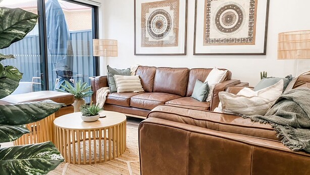 A modern living room with a brown leather sofa, a round wooden coffee table, and two framed abstract artworks on the wall. - Coast and Roast - Sunny Shell Cove Escape (Shell Cove)