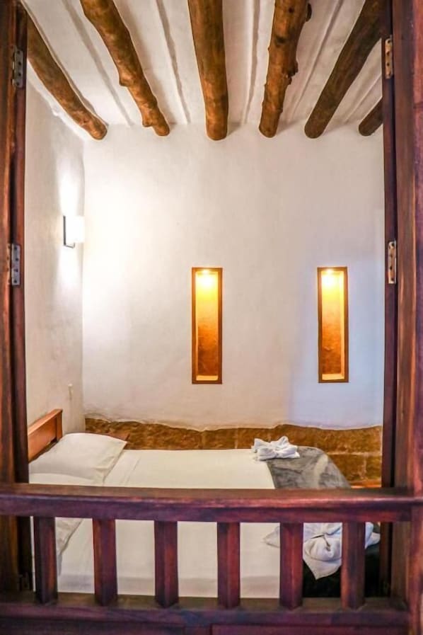 Une pièce avec un plafond en bois, deux lampes fixées au mur, un lit avec une literie blanche et une balustrade en bois.