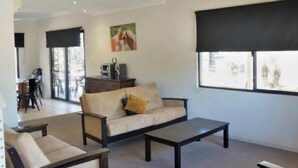 A spacious living area with a sofa, coffee table, and chairs. There is a large window with blinds, a painting on the wall, and a door leading to another room.