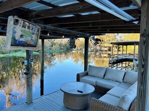 A covered outdoor seating area with a view of a calm body of water, a television mounted on a wooden post, and a fan on the ceiling. - Sleeps 13, theater, waterfront, close to springs and forest. Old Florida (Astor)