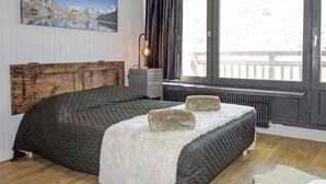 A bedroom with a wooden headboard, a bed with a grey quilt, a white fluffy throw, and a painting of a mountain landscape. - Le Roc de Peclet by Interhome (Val Thorens)