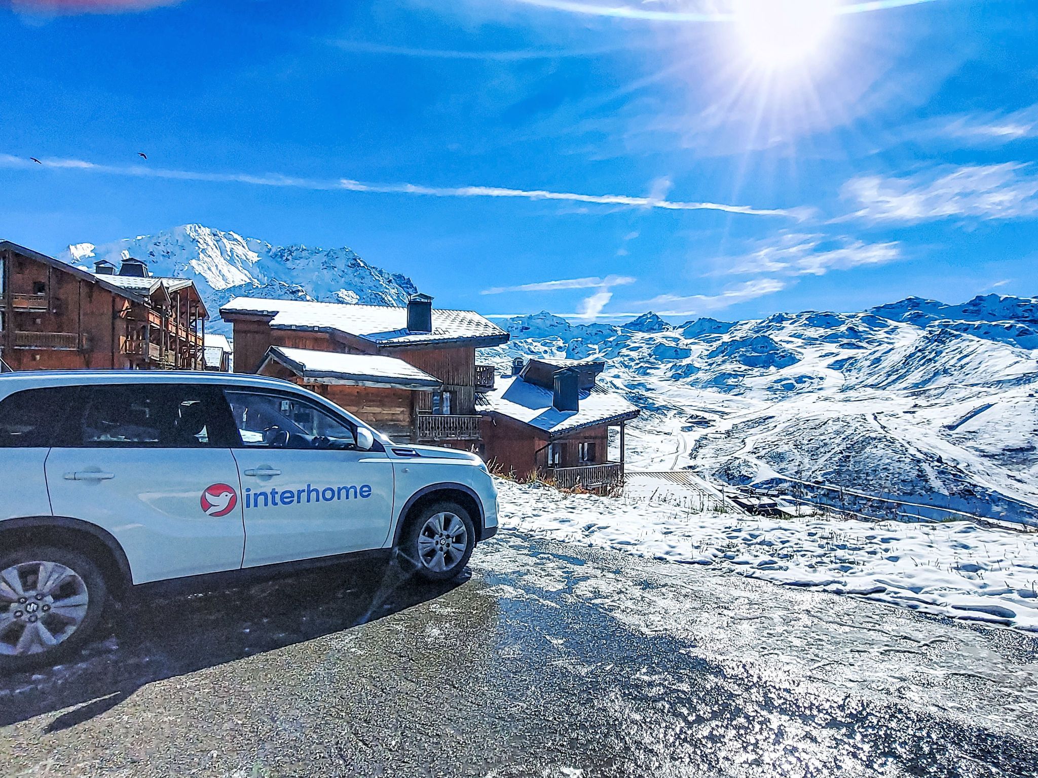 A white Interhome vehicle parked in a snowy mountain area with wooden buildings and snow-covered mountains in the background.