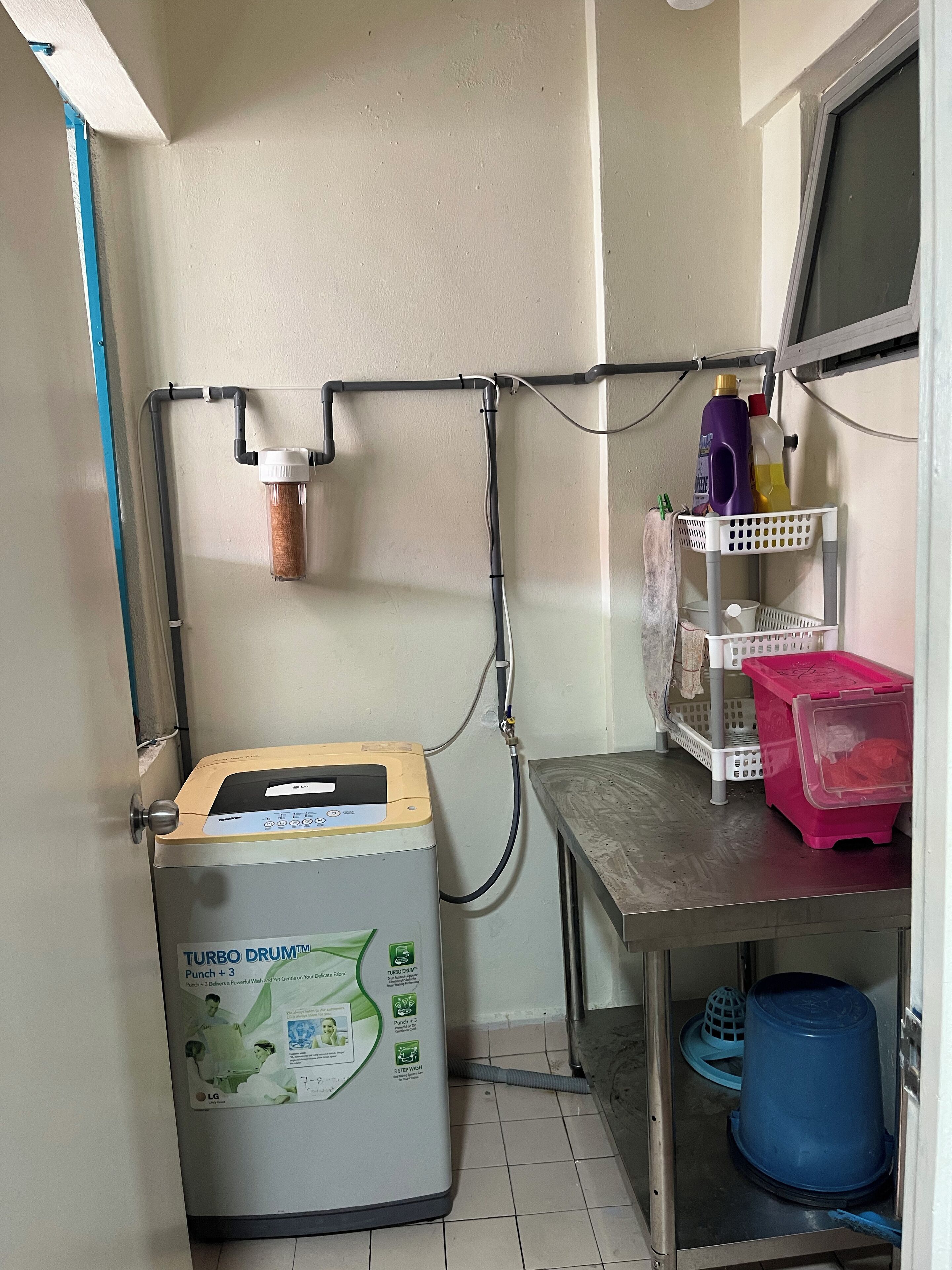 Una lavanderia funzionale con una lavatrice Turbo Drum, uno scaldabagno e un piccolo tavolo con cesti portaoggetti e prodotti per la pulizia.