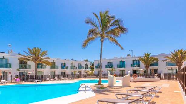 A hotel pool area with multiple lounge chairs, palm trees, and a clear blue sky.