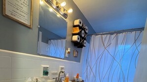 Un bagno con lavandino, servizi igienici e uno specchio. Si vedono rotoli di carta igienica su un porta carta, una piccola pianta sul lavandino e una tenda da doccia con motivo ondulato.