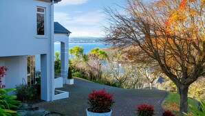 A modern house with a paved driveway leading to a white entrance, surrounded by autumn foliage and a view of a body of water. - ACACIA WATERS (Taupo)