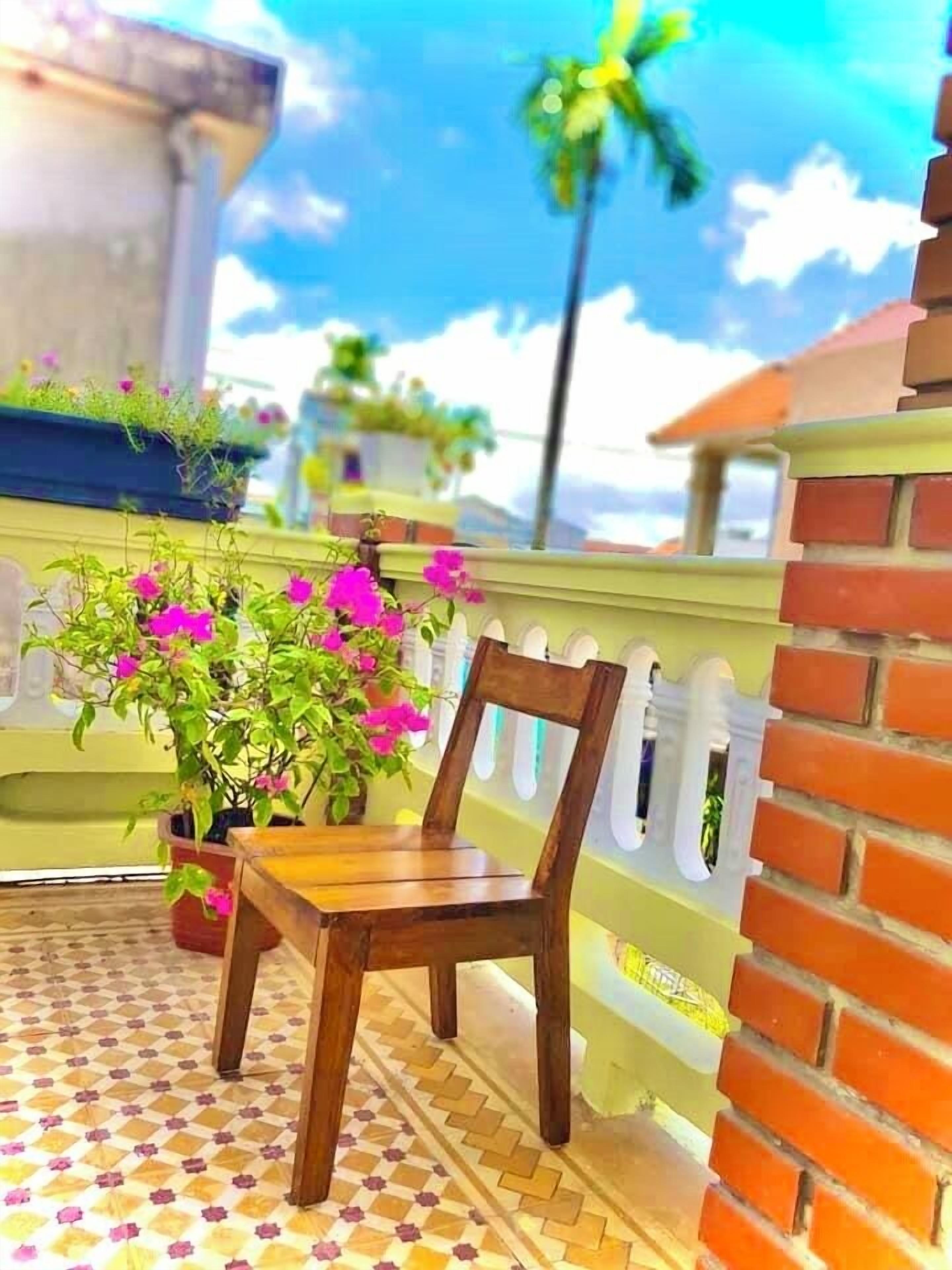 Una silla de madera en un balcón con piso de baldosas, rodeada de plantas en macetas y vista al cielo.