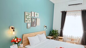 A hotel room with a bed, a nightstand with a welcome sign, a small table with a plant, and a window with curtains. - Hidden Hue Homestay (Hue)