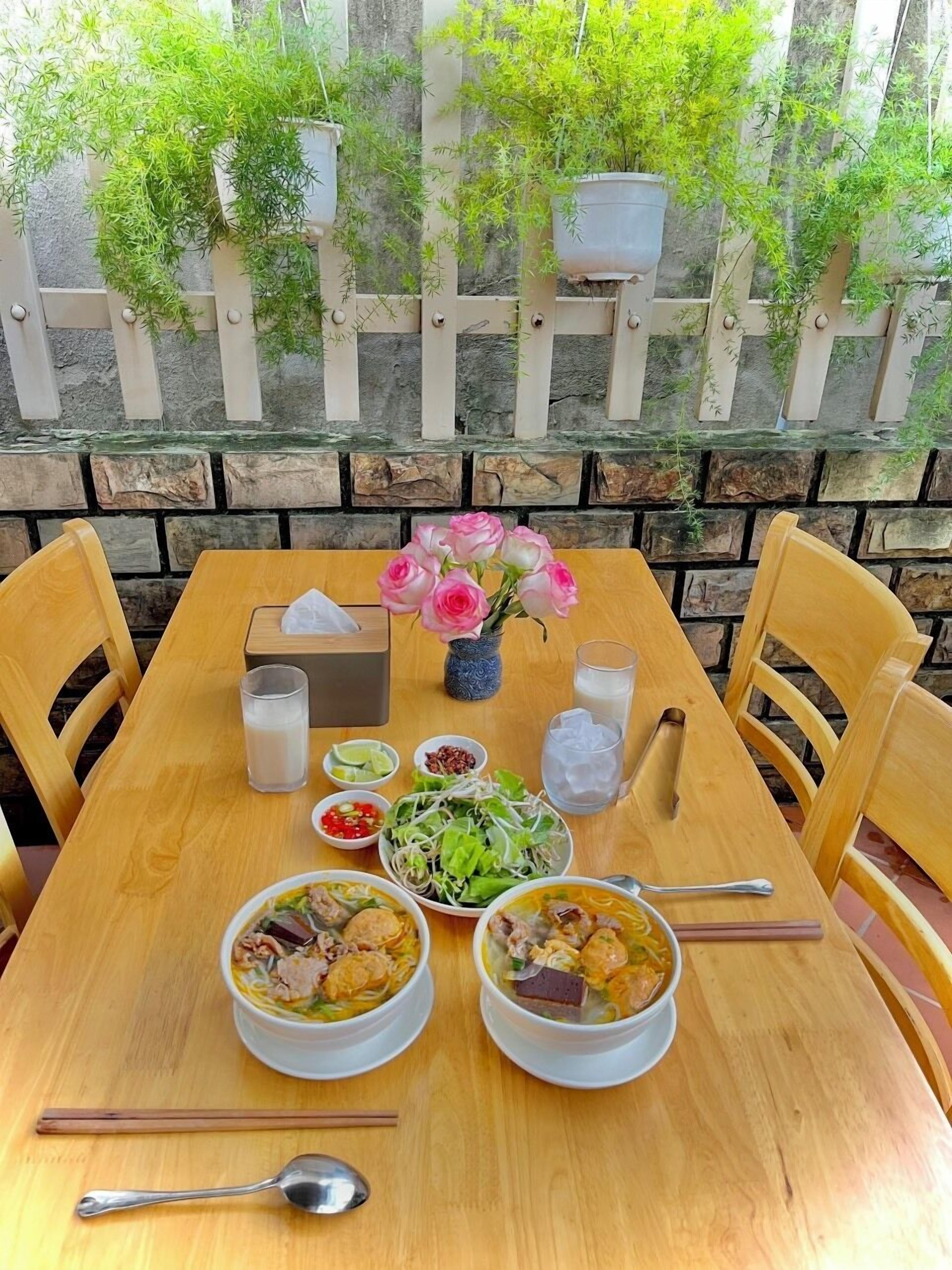 A wooden table set with two bowls of soup, a bowl of salad, a vase of pink roses, and various condiments.