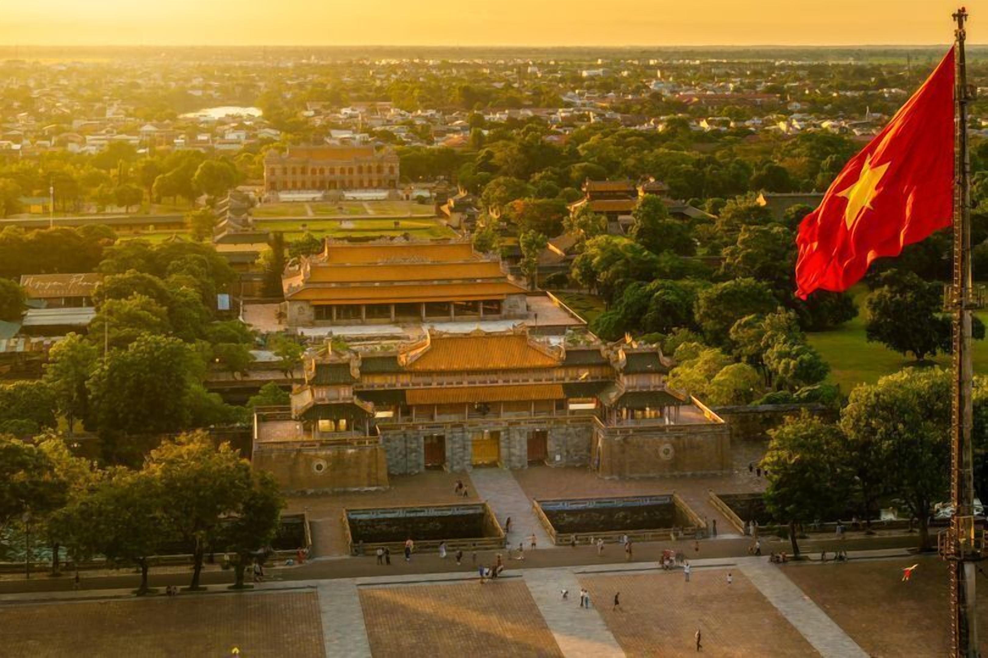 A traditional Vietnamese temple complex with a prominent flag, surrounded by lush greenery and a clear view of the cityscape in the background.