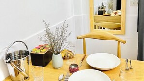 A wooden dining table with two white plates, a metal bucket, a glass, a spoon, a fork, a knife, a vase with dried flowers, and a small bowl of lemons. - Hidden Hue Homestay (Hue)