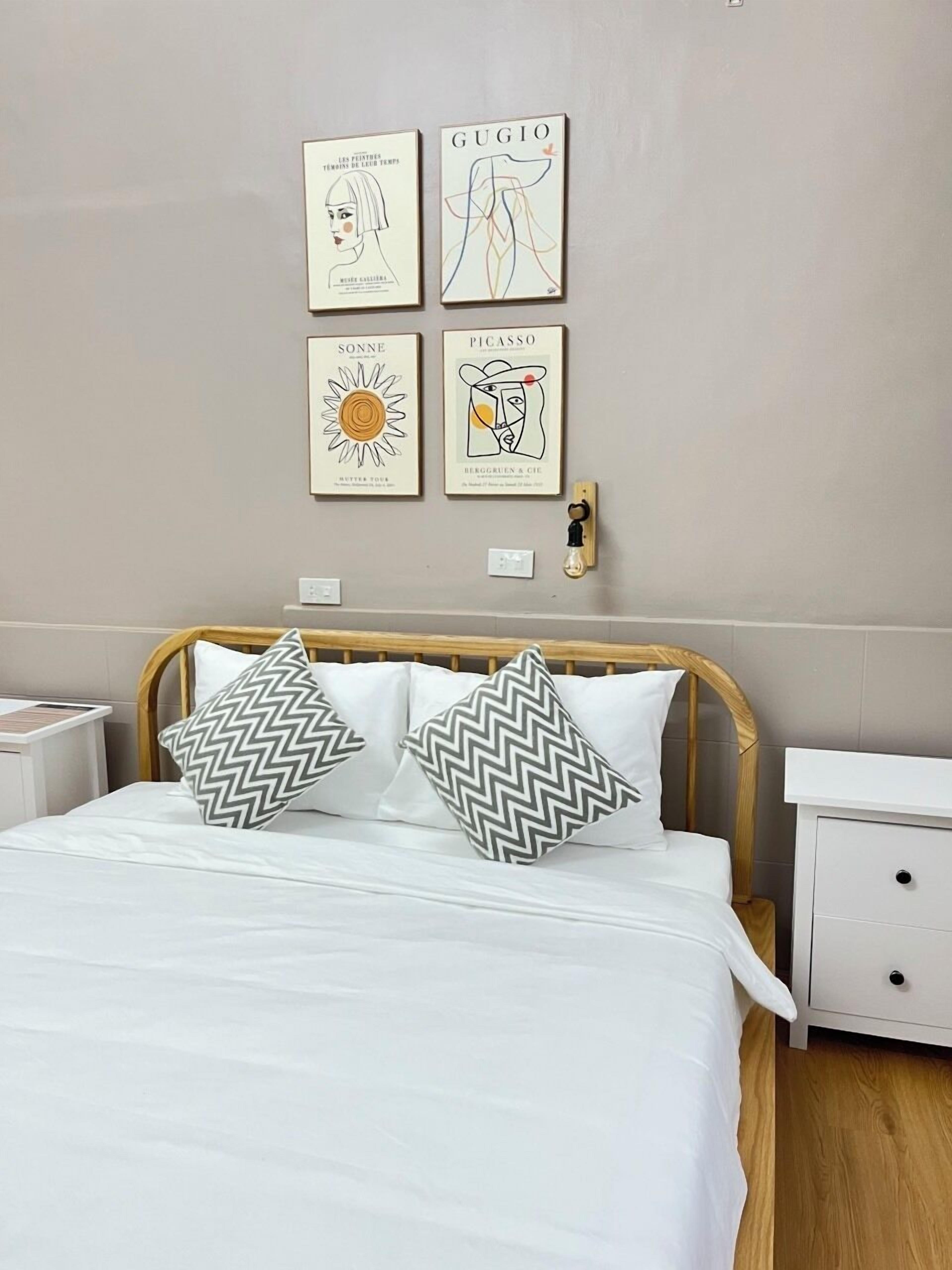 Una cama bien hecha con dos almohadones estampados, un cabecero de madera y una mesita de noche con una lámpara. Cuatro obras de arte enmarcadas están colgadas en la pared sobre la cama.
