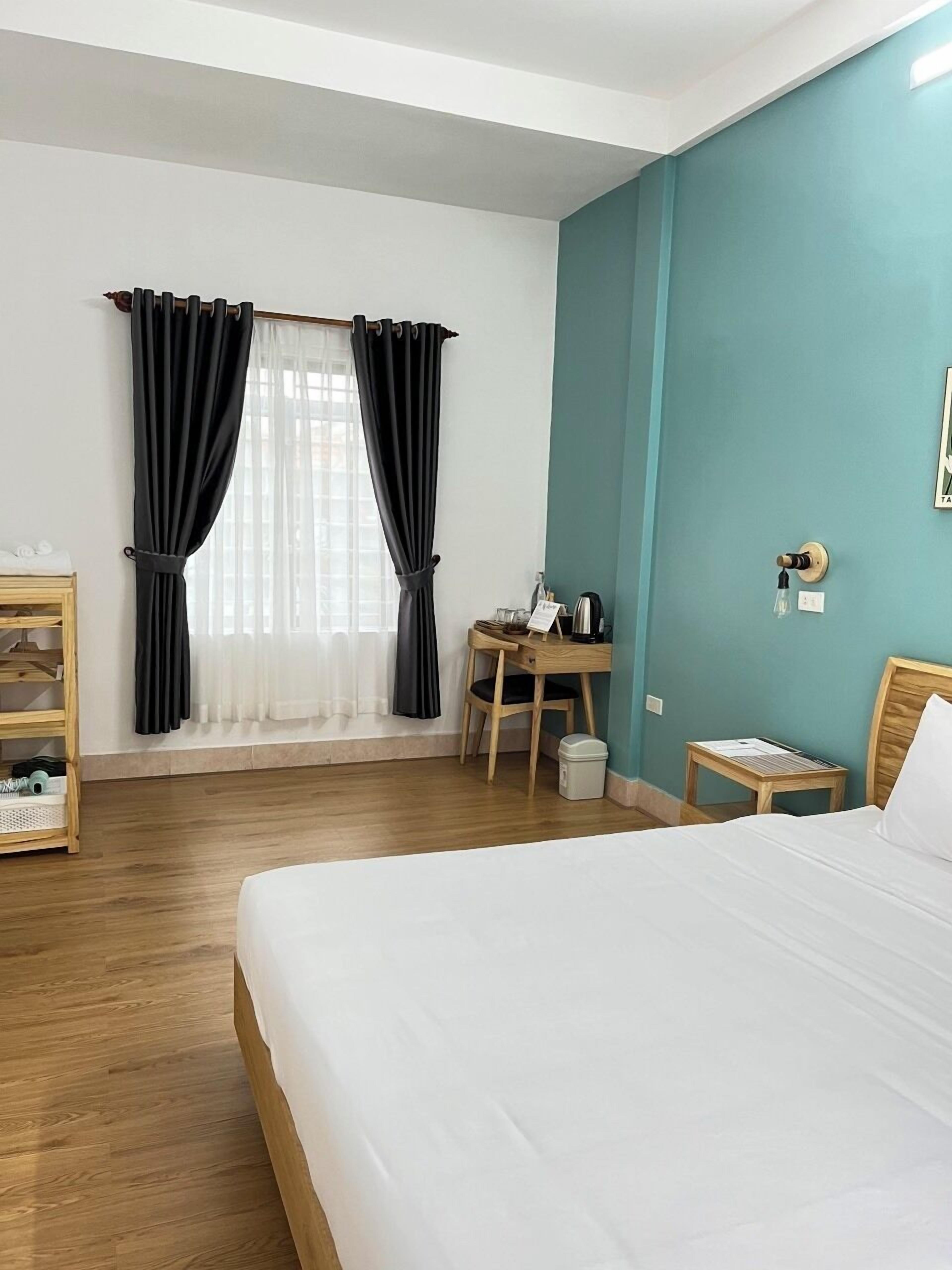 A hotel room with a bed, a desk, a chair, a small table, a shelf, and a window with curtains.