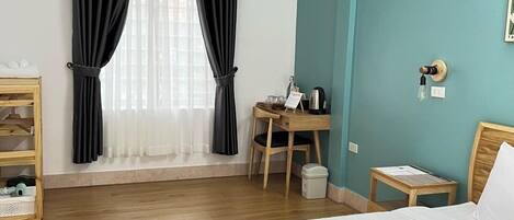 A hotel room with a bed, a desk, a chair, a small table, a shelf, and a window with curtains.