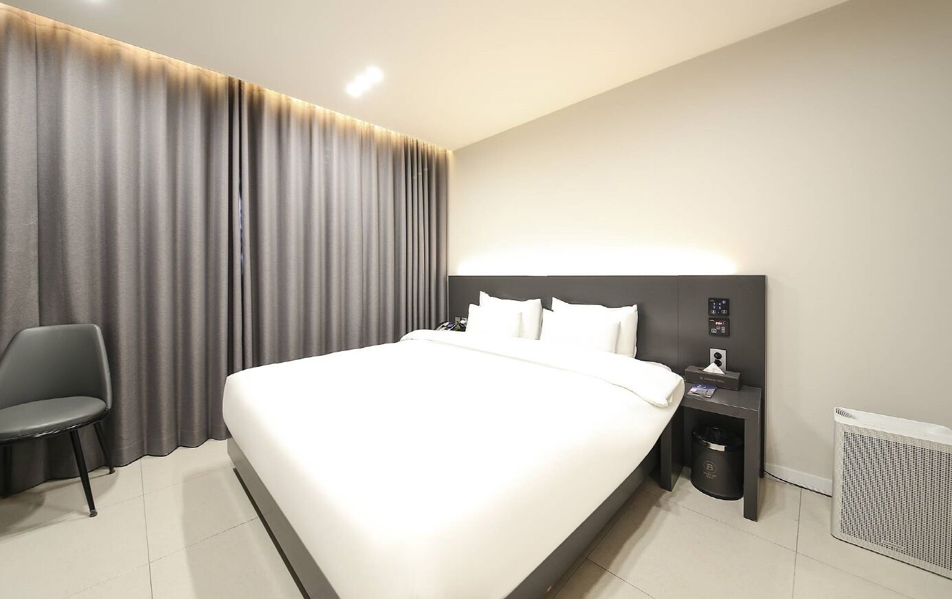 A modern hotel room with a large bed, a bedside table with a lamp, a chair, and a waste bin.