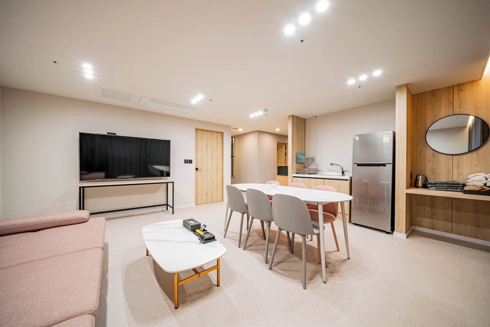 A modern living room with a flat-screen TV, a sofa, a dining table with chairs, a kitchen area with a refrigerator, and a wooden accent wall with a mirror.