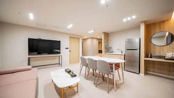 A modern living room with a flat-screen TV, a sofa, a dining table with chairs, a kitchen area with a refrigerator, and a wooden accent wall with a mirror.