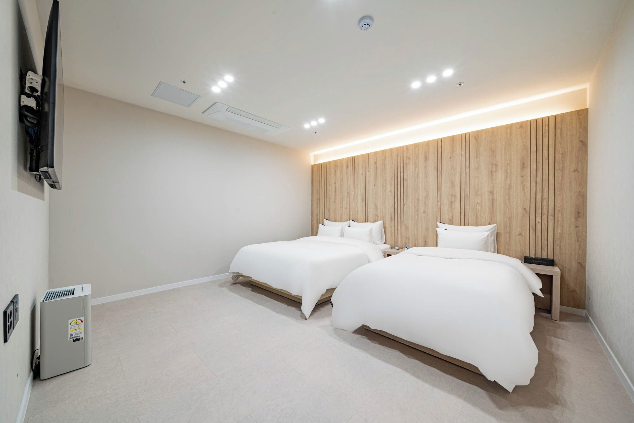 Una habitación de hotel moderna con dos camas, un cabecero de madera, un televisor de pantalla plana en la pared y una mesita de noche con una lámpara.