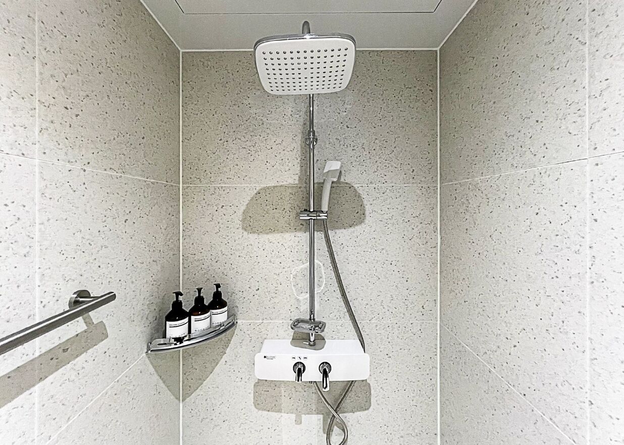A shower with a handheld showerhead, a wall-mounted control panel, and a shelf with toiletries.
