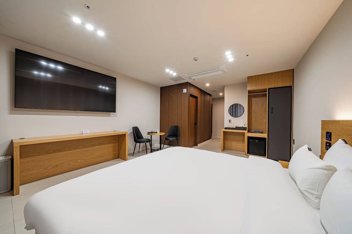 A modern hotel room with a large bed, a wooden desk, a flat-screen TV, a wardrobe, and a small seating area.