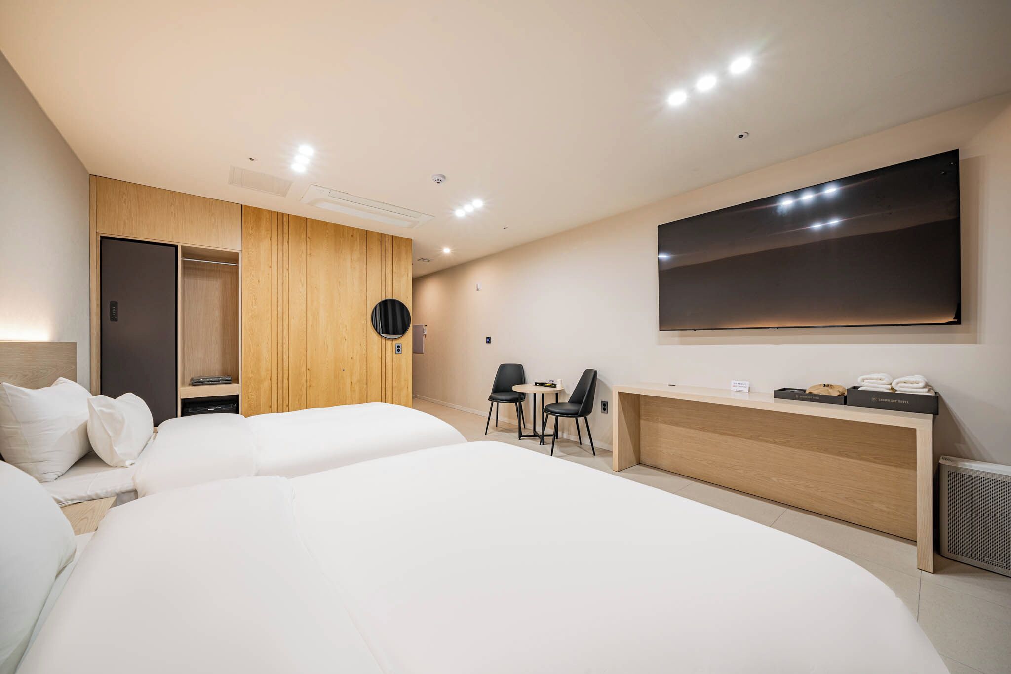 A modern hotel room with a large bed, a desk, two chairs, a wardrobe, and a large TV on the wall.