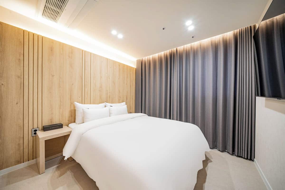 A modern hotel room with a wooden panel wall, a large bed with white bedding, a bedside table with a remote control, and a large window with grey curtains.