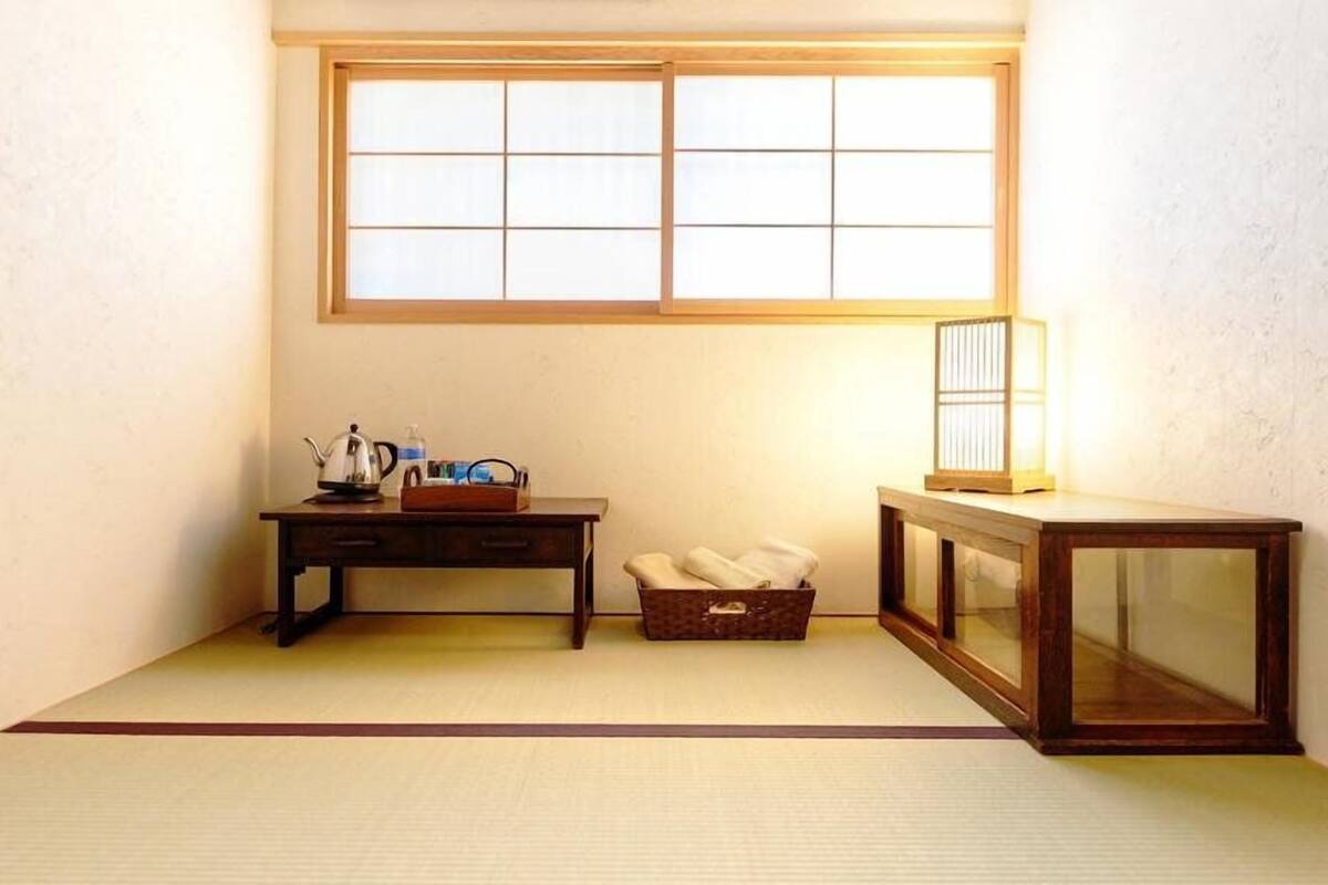 Een kamer met tatami vloerbedekking, een laag houten bureau met waterkoker en waterfles, een klein houten kastje en een raam met uitzicht.