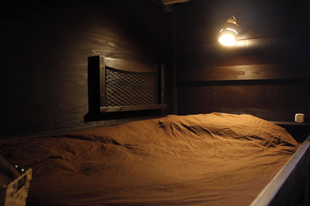 Een donkere kamer met een bed, een raam met hor en een wandlamp.