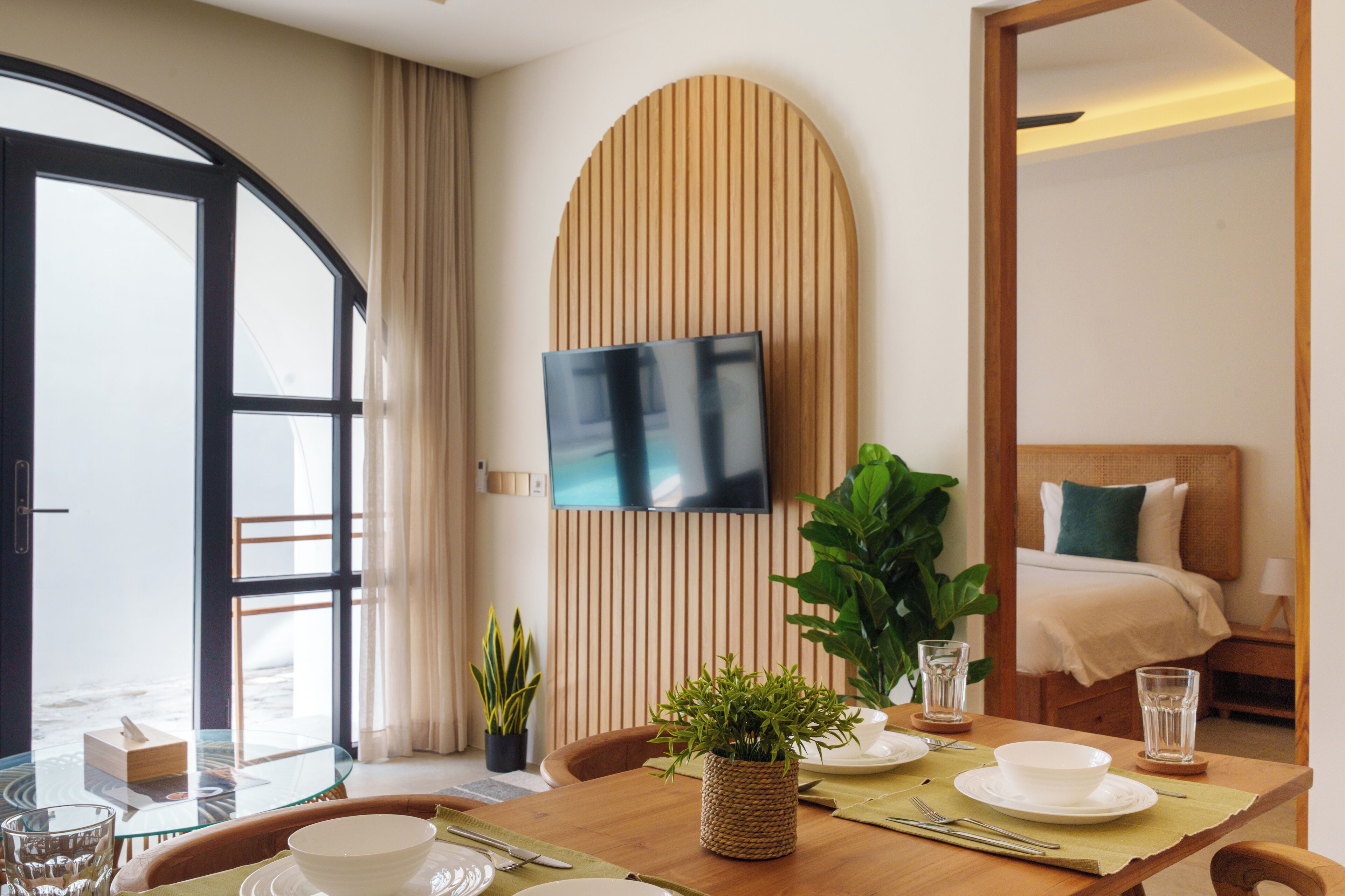 A modern hotel room with a dining area, a bed with a green pillow, a TV mounted on a wooden wall, and a large window with curtains.